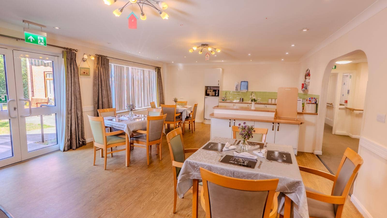 The image depicts a bright and inviting dining area. The space features several tables set with white tablecloths and neatly arranged cutlery, suggesting a welcoming atmosphere for meals. Soft lighting from modern fixtures illuminates the room, enhancing