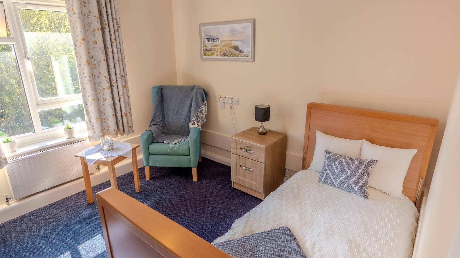 The image depicts a cozy bedroom with a warm and inviting atmosphere. The room features a single bed with a light-colored blanket and decorative pillows. Next to the bed is a small bedside table with a lamp. A comfortable green armchair is positioned in o