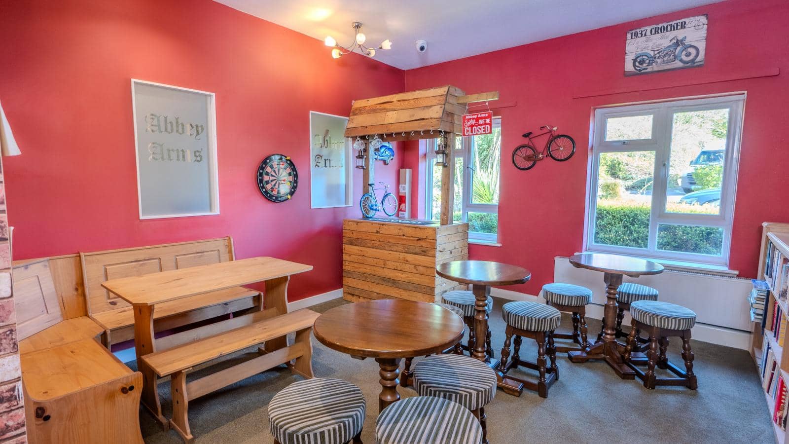 The image depicts a cozy interior of a pub or café with a warm, inviting atmosphere. The walls are painted a vibrant shade of red, complemented by wooden accents. There are several wooden tables, including a larger communal table and smaller round tables